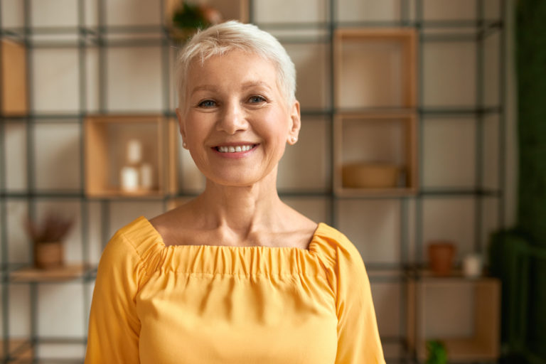 Portrait of joyful stylish middle aged woman with short haircut posing indoors expressing positive emotions, looking at camera with broad happy smile, wearing yellow summer dress